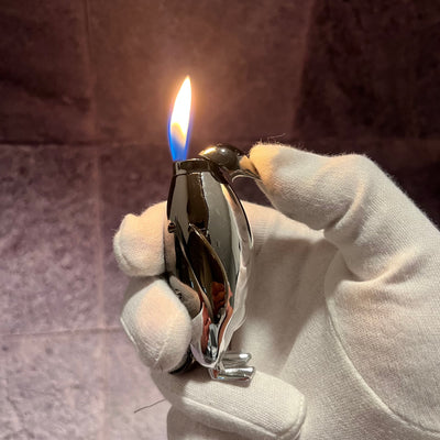 Penguin Flame Lighter – chrome silver refillable butane torch lighter shaped like a penguin, producing a powerful blue flame while held in a gloved hand.